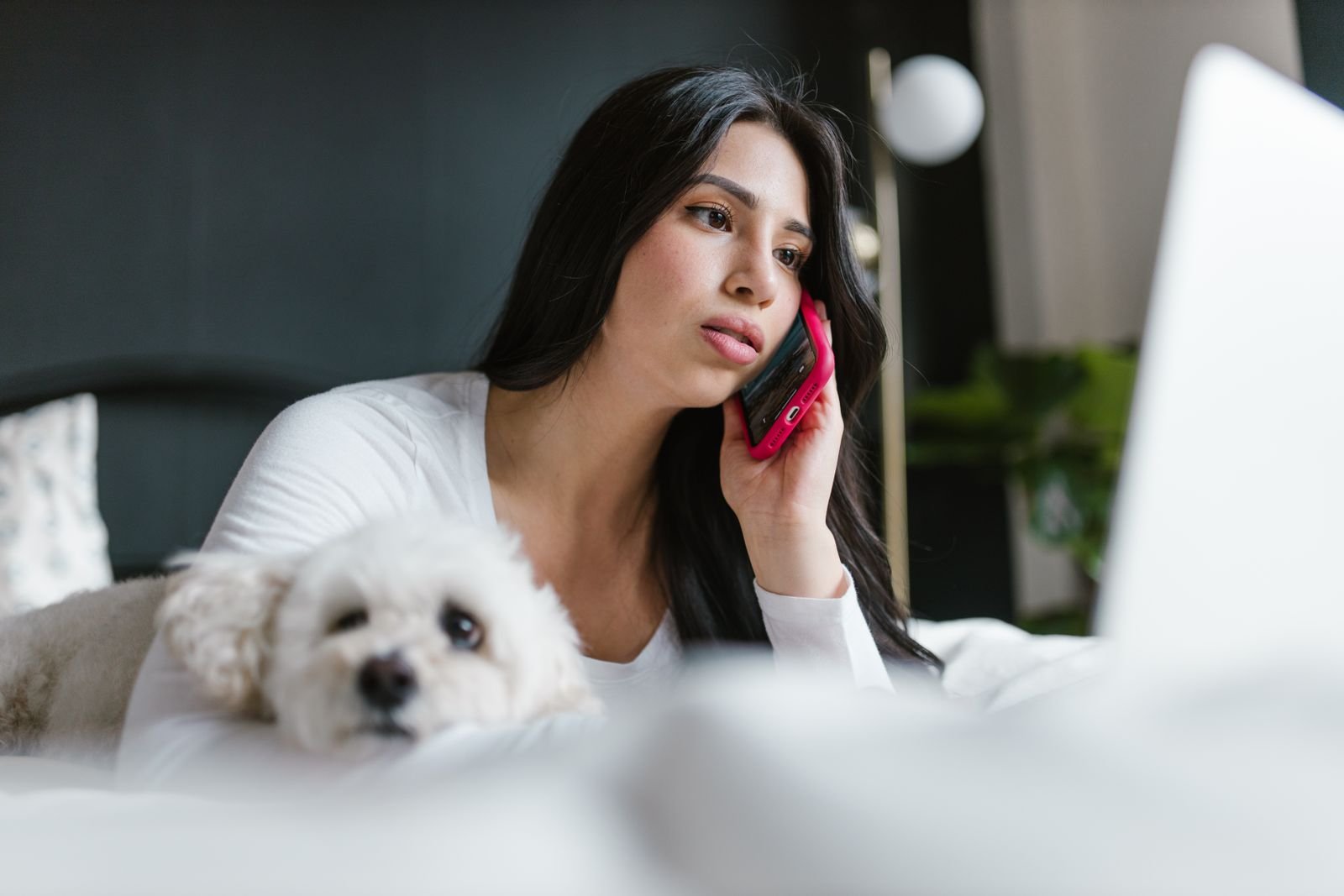 Woman talking on the phone at home with her small white dog resting beside her, calling for advice after her dog ate chocolate